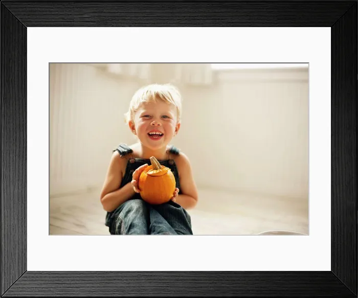 Framed Print : Young boy holds a small Jack O'Lantern on his knees as he sits on a floor and poses for a portrait Framed Print : Young boy holds a small Jack O'Lantern on his knees as he sits on a floor and poses for a portrait