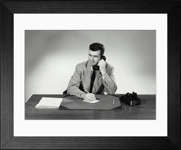 Framed Print : Vintage Photograph. Man sits at desk taking notes on small card smiling he talks on phone