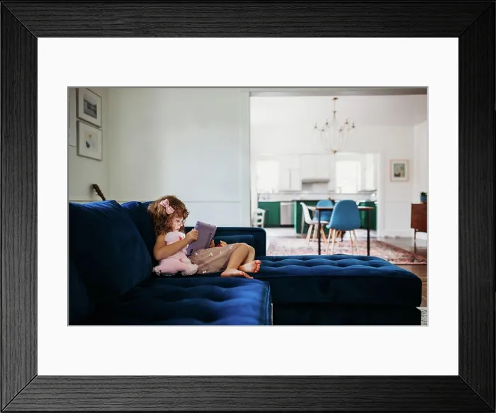 Framed Print : Young girl sitting on couch in modern living room learning on tablet Framed Print : Young girl sitting on couch in modern living room learning on tablet