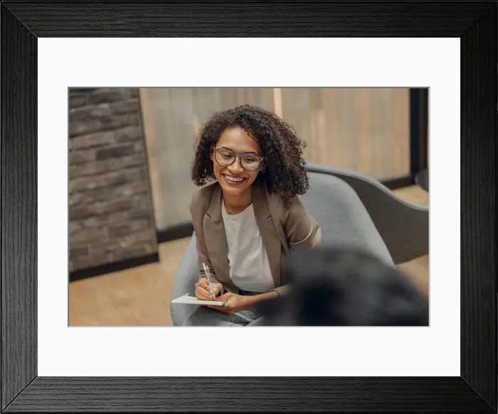 Framed Print : Smiling woman writing something down while sitting with colleague in modern office Framed Print : Smiling woman writing something down while sitting with colleague in modern office