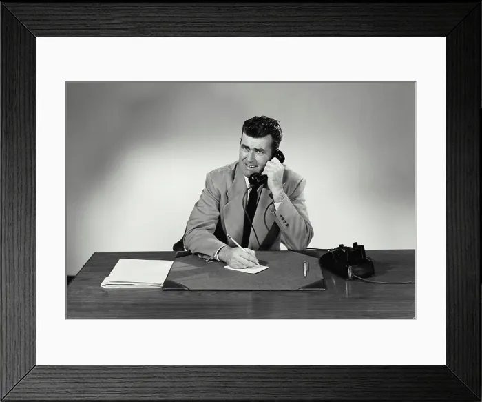 Framed Print : Vintage Photograph. Man sits at desk taking notes he smiles with a concerned look on his face he talks on phone Framed Print : Vintage Photograph. Man sits at desk taking notes he smiles with a concerned look on his face he talks on phone