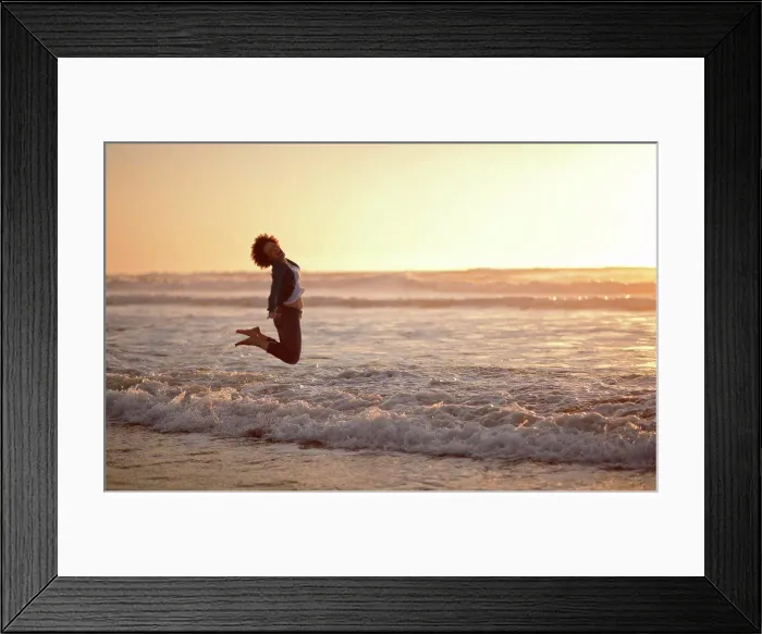 Framed Print : Portrait of a laughing woman leaping into the air while standing at the edge of the sea on a beach