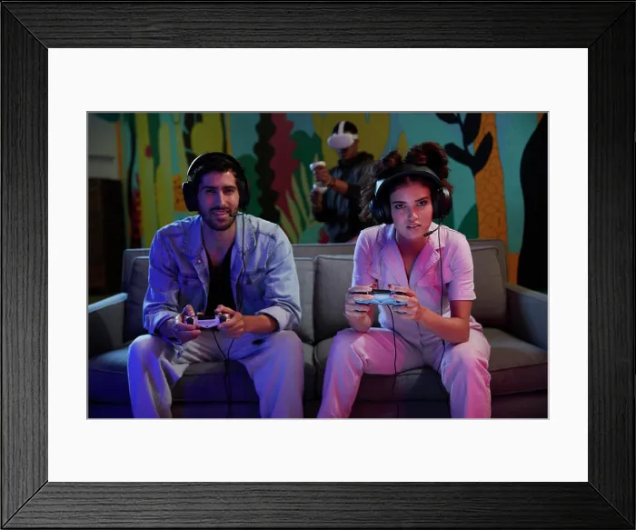 Framed Print : Two friends enjoy a competitive video game while another person experiences virtual reality in the background