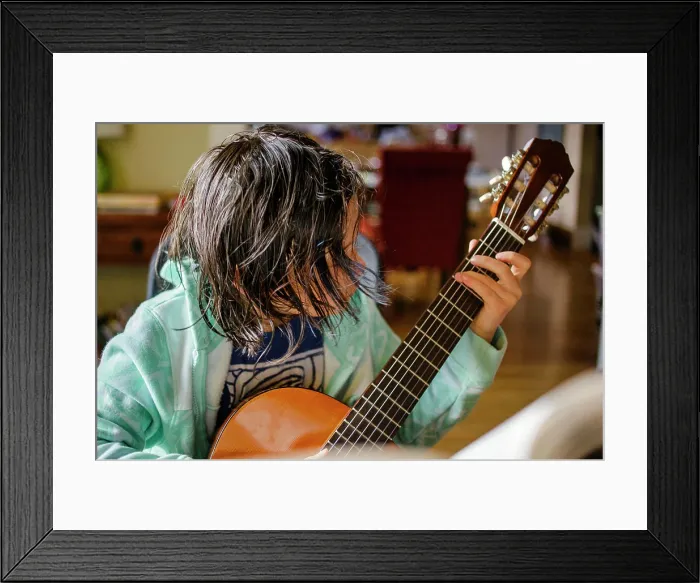 Framed Print : A child with hair hanging in his face practices classical guitar