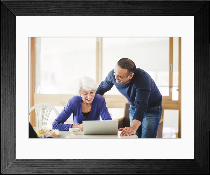 Framed Print : Cheerful mature man standing by mother looking at laptop on table in nursing home Framed Print : Cheerful mature man standing by mother looking at laptop on table in nursing home
