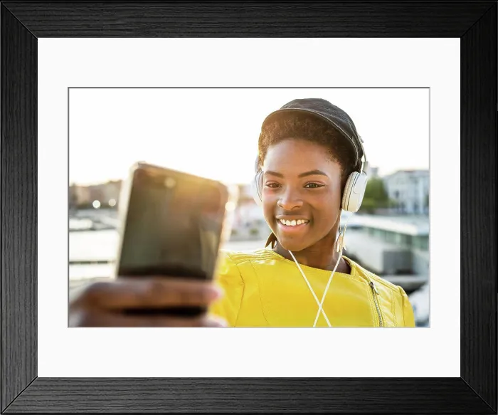 Framed Print : African American woman in stylish bright jacket using mobile phone and listen to music on headphones
