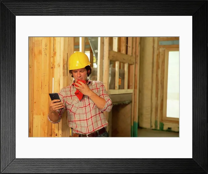 Framed Print : Construction worker on job site feeling ill, sneezing, using mobile phone to check in with tele-medicine doctor Framed Print : Construction worker on job site feeling ill, sneezing, using mobile phone to check in with tele-medicine doctor