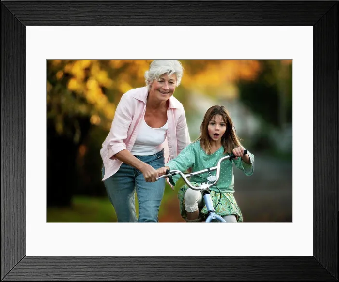 Framed Print : Little girl learning to ride her bicycle with the help of her grandmother Framed Print : Little girl learning to ride her bicycle with the help of her grandmother