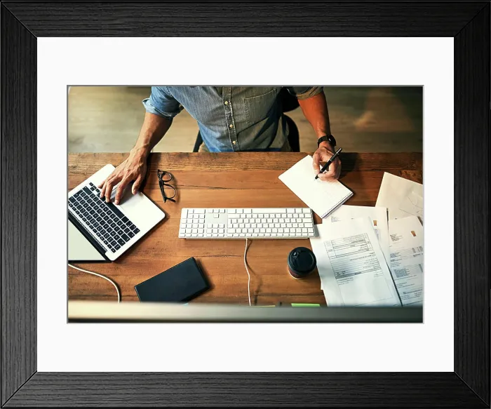 Framed Print : High angle shot of a businessman using a computer and writing notes during a late night at work Framed Print : High angle shot of a businessman using a computer and writing notes during a late night at work