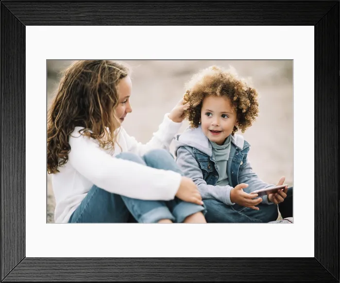 Framed Print : Smiling casual curly child resting on ground with crossed legs holding mobile phone while content girl sitting clasping
