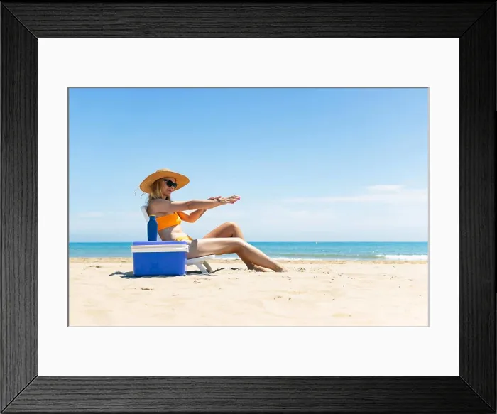Framed Print : a tanned girl in a swimsuit, hat and glasses sits on a sun lounger smears sunscreen on her hands