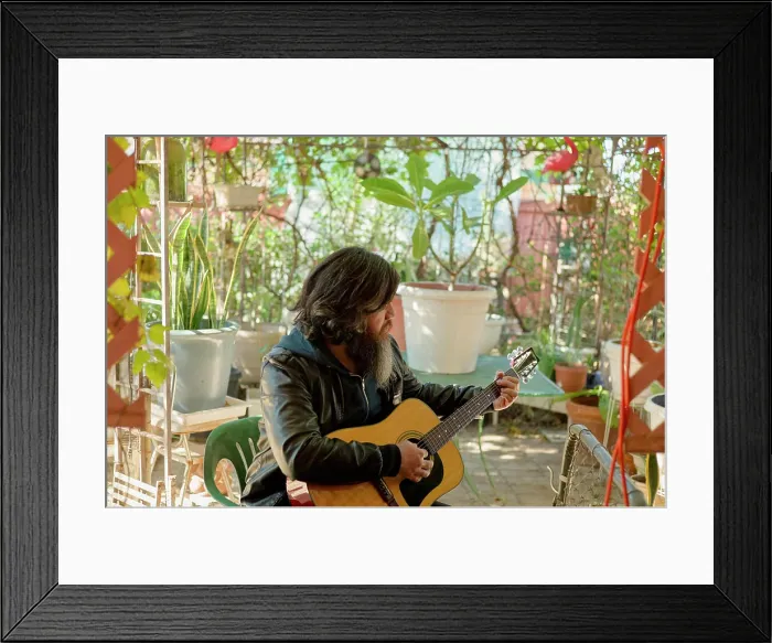 Framed Print : A bearded man with long hair plays an acoustic guitar in a cozy garden surrounded by potted plants and decorative items