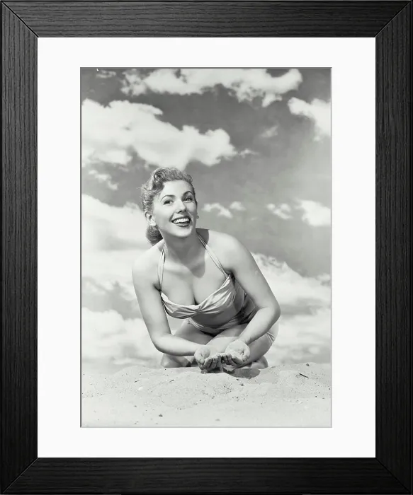 Framed Print : Vintage photograph. Young smiling woman in swimsuit kneeling in sand with sky background