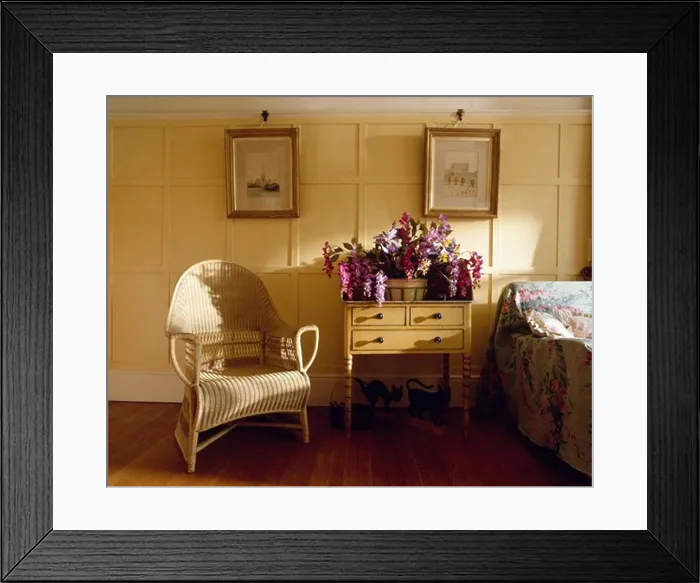 Framed Print : Pictures on cream panelled walls of country bedroom with wicker armchair and flowers on small antique table