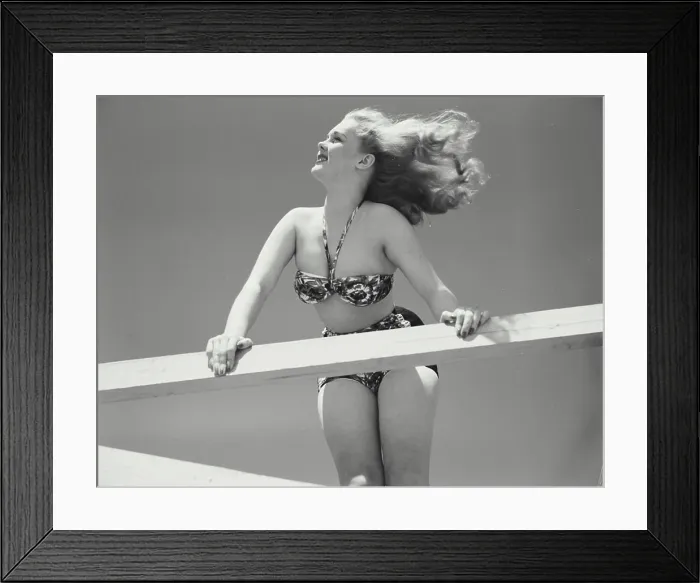 Framed Print : Woman in bikini leaning over railing of deck with hair blowing in wind