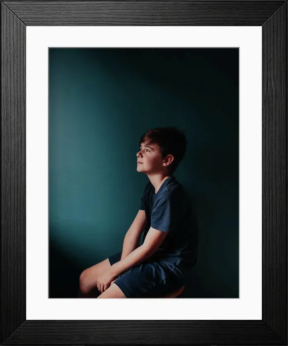 Framed Print : Thoughtful young boy sitting on a stool against a dark blue wall