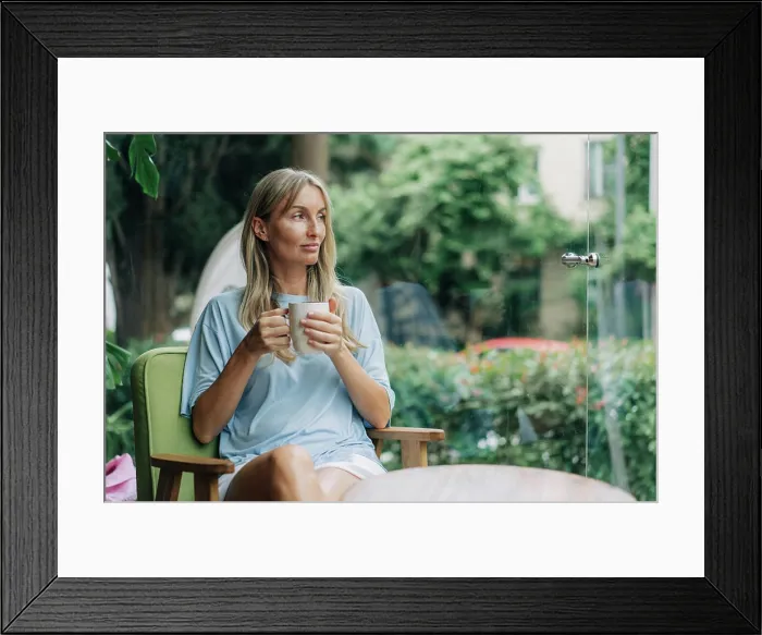 Framed Print : Eine junge Frau sitzt in einem Café in der Nähe des Fensters und trinkt ihren Morgenkaffee Framed Print : Eine junge Frau sitzt in einem Café in der Nähe des Fensters und trinkt ihren Morgenkaffee