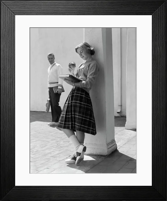 Framed Print : 1950S 1960S Teen School Girl Plaid Skirt Leaning Against Column Eating Apple Reading Book As Boy Walks By In Background Framed Print : 1950S 1960S Teen School Girl Plaid Skirt Leaning Against Column Eating Apple Reading Book As Boy Walks By In Background