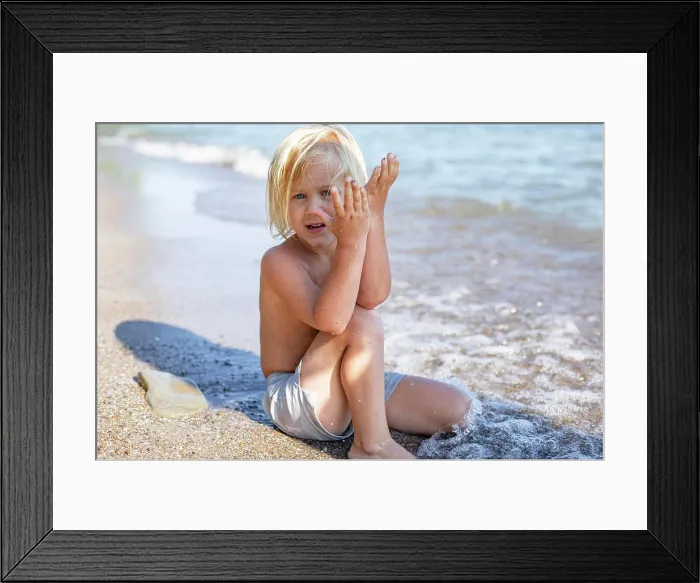 Framed Print : A young child sitting on a sandy beach covering one side of their face with sand-covered hands Framed Print : A young child sitting on a sandy beach covering one side of their face with sand-covered hands
