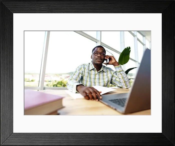 Framed Print : African man entrepreneur talking on cellphone and working on laptop at office Framed Print : African man entrepreneur talking on cellphone and working on laptop at office