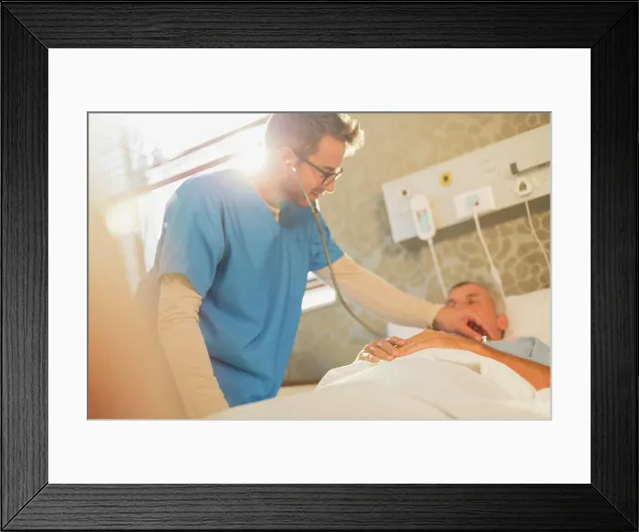 Framed Print : Male nurse using stethoscope on patient in hospital bed