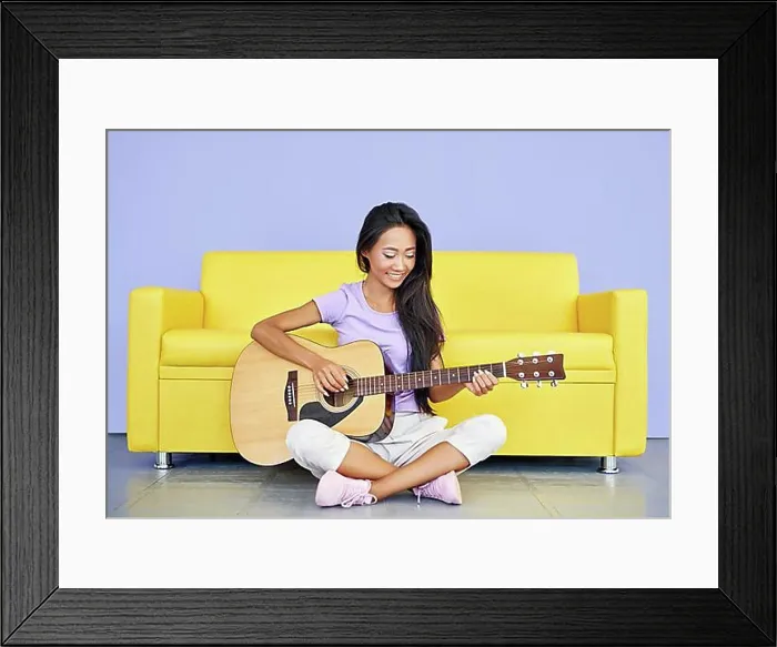 Framed Print : Smiling pretty female playing guitar sitting on floor leaning sofa. Creative, hobby, music concept