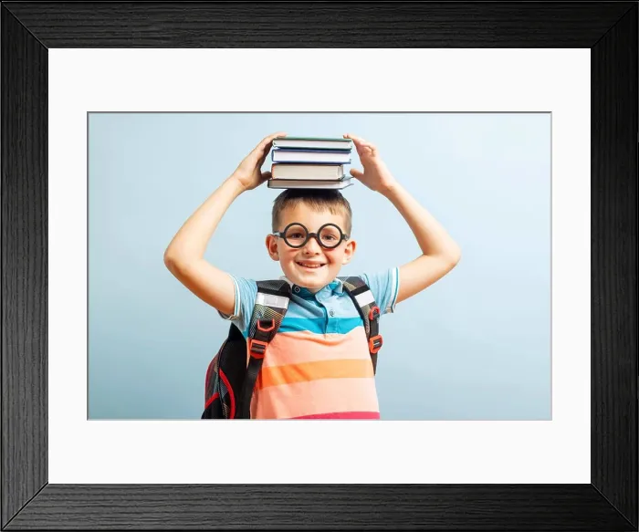 Framed Print : Funny smiling school boy in glasses holds books on his head on blue background Framed Print : Funny smiling school boy in glasses holds books on his head on blue background