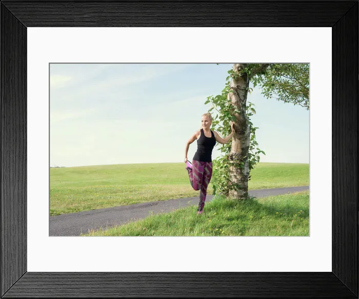 Framed Print : Fit female athlete stretching leg near tree during training in meadow