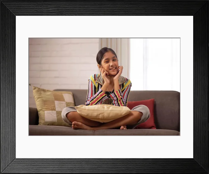 Framed Print : Cheerful girl sitting on sofa and looking at camera with hands on cheek Framed Print : Cheerful girl sitting on sofa and looking at camera with hands on cheek