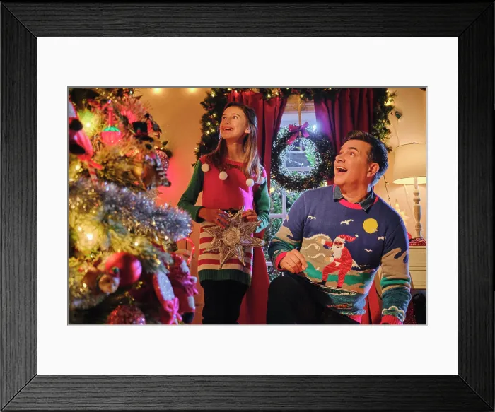 Framed Print : Couple decorating Christmas tree together wearing festive holiday sweaters in colorfully lit living room