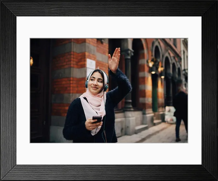 Framed Print : Smiling teenage girl waving hand while listening to headphones on footpath in city Framed Print : Smiling teenage girl waving hand while listening to headphones on footpath in city