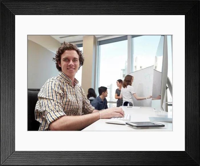 Framed Print : Young man using computer in office looking towards camera, co-workers having discussion in background Framed Print : Young man using computer in office looking towards camera, co-workers having discussion in background