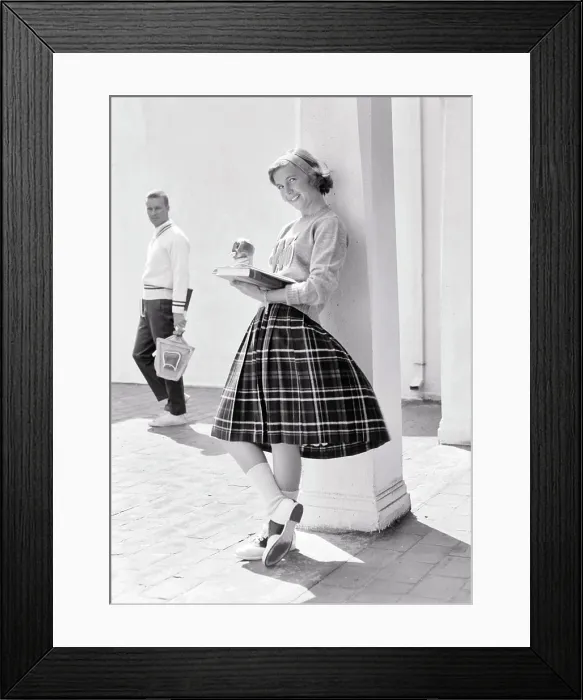 Framed Print : 1950s 1960s TEEN GIRL LEANING AGAINST COLUMN EATING APPLE READING BOOK LOOKING AT CAMERA SMILING AS POTENTIAL BOYFRIEND