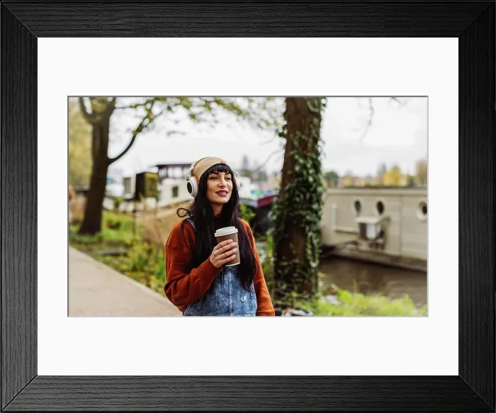 Framed Print : Smiling woman holding coffee cup and listening to music through wireless headphones
