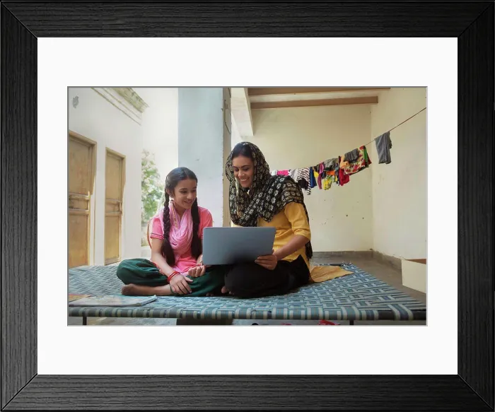 Framed Print : Happy village woman sitting with her daughter on a cot at home and operating a laptop computer