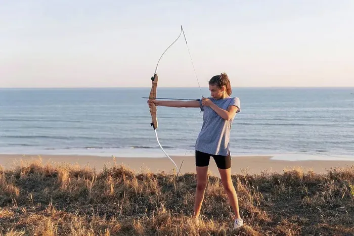 Framed Print : Archeress aiming with bow and arrow while standing on grass at beach
