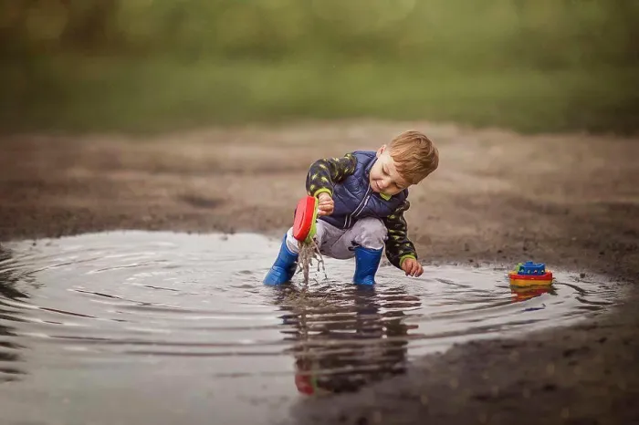 Framed Print : Boy in rain boots crouching in puddle and playing with ships Framed Print : Boy in rain boots crouching in puddle and playing with ships
