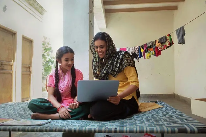 Framed Print : Happy village woman sitting with her daughter on a cot at home and operating a laptop computer
