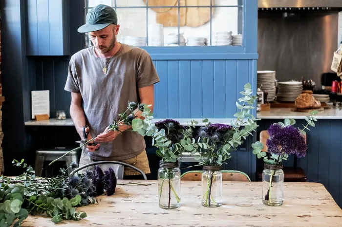 Framed Print : Man wearing baseball cap standing at a table, cutting thistles for flower arrangements in large glass jars
