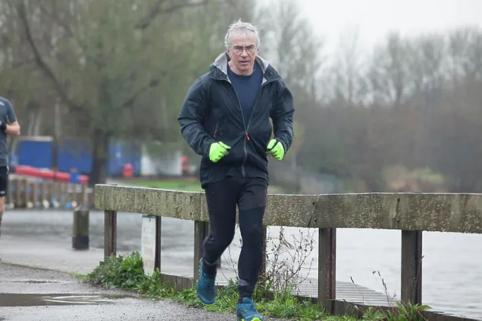 Framed Print : A mature man jogging along a waterside path on an overcast day, wearing a black jacket and bright green gloves