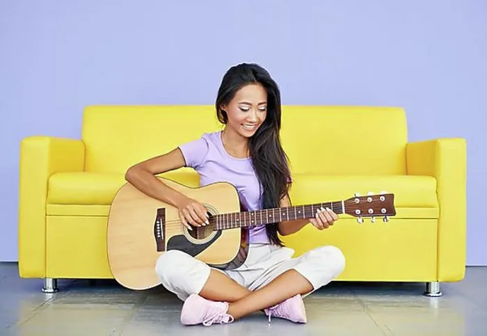 Framed Print : Smiling pretty female playing guitar sitting on floor leaning sofa. Creative, hobby, music concept
