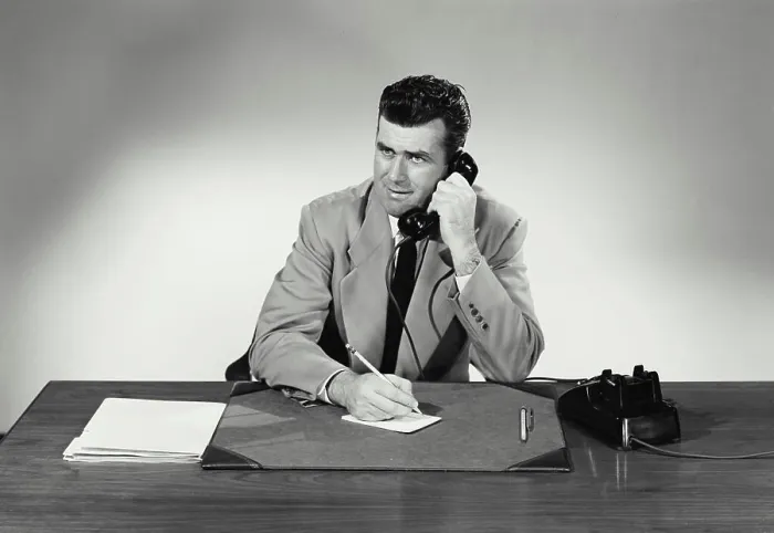 Framed Print : Vintage Photograph. Man sits at desk taking notes on small card smiling he talks on phone