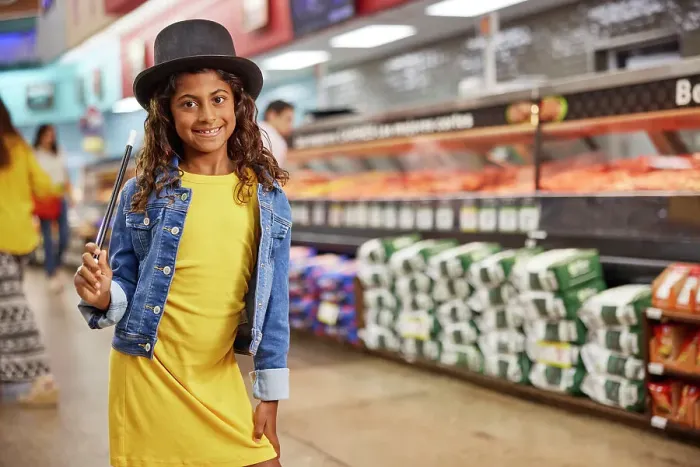 Framed Print : Young girl with magicians top hat and wand standing in isle of grocery store, meat market in the background