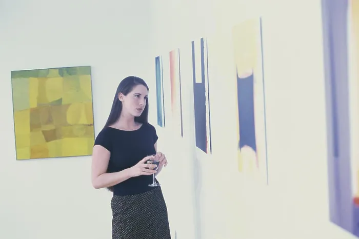 Framed Print : Young woman holding a glass looking at paintings on a wall Framed Print : Young woman holding a glass looking at paintings on a wall