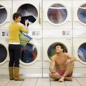 Man in boxers sleeping at Laundromat with woman removing clothing from dryer