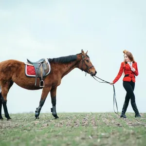 red-haired jockey girl in a red cardigan and black high boots with a horse for a walk