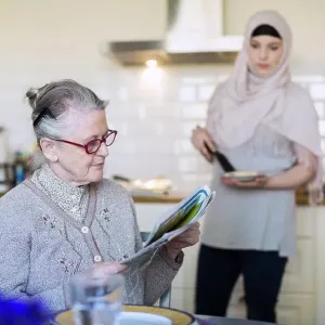 Senior woman reading newspaper with female home caregiver preparing food in kitchen Senior woman reading newspaper with female home caregiver preparing food in kitchen