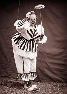 Circus Prints: 1930s BIG CLOWN UNDER TINY UMBRELLA TIPPING HIS SMALL HAT LOOKING AT CAMERA WEARING CLOWN SHOES OVERSTUFFED STRIPED