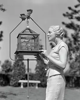 Styling Prints: 1930s BLONDE WOMAN WITH MARCELLED HAIR STYLE OUTDOOR IN GARDEN WHISTLING OR TALKING TO A PET BIRD IN A BIRDCAGE