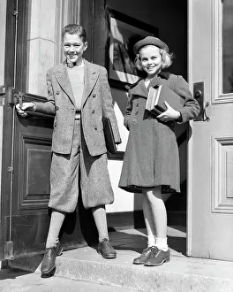 Expressions Prints: 1930s LOOKING AT CAMERA SMILING PRE-TEENAGER BOY IN KNICKERS HOLDING DOOR FOR GIRL IN HAT ENTERING LEAVING SCHOOL